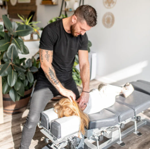 Dr. Dustin McIver, a chiropractor in Grand Rapids, wearing a black t-shirt and dark pants, performs a neck adjustment on a female patient lying face down on a chiropractic adjustment table. The clinic space is bright, with large plants in the background.